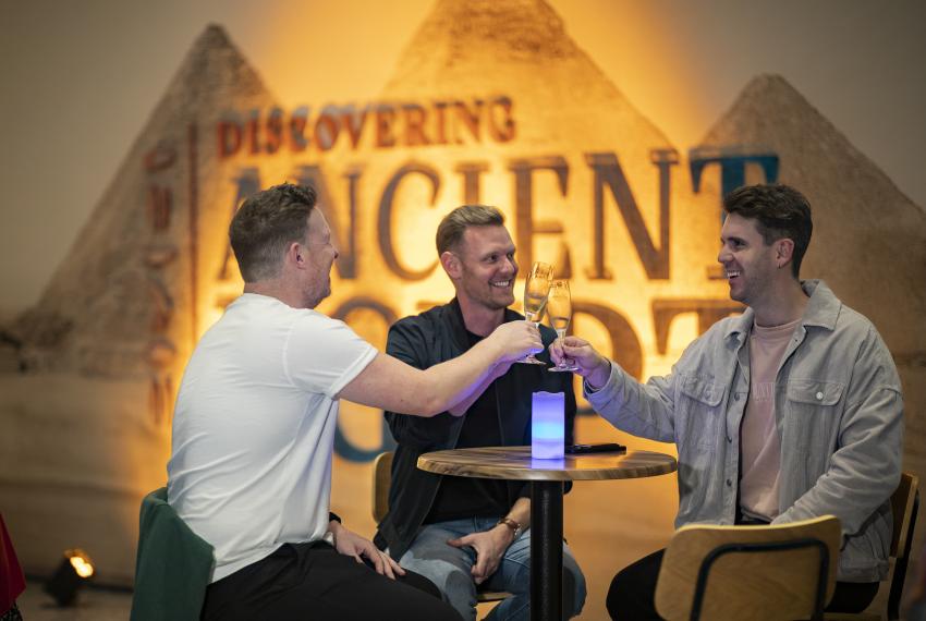 Three people smiling at each other and clinking slender champagne glasses while seated at a high bar table in front of an impressively lit image of three pyramids with the words Discovering Ancient Egypt printed over the top