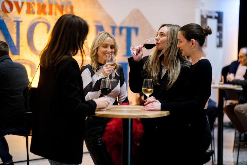 Four people standing around a cocktail bar with a wooden top laughing, and holding or drinking glasses of red and white wine. Behind them the entrance to the Discovering Ancient Egypt gallery is brightly lit