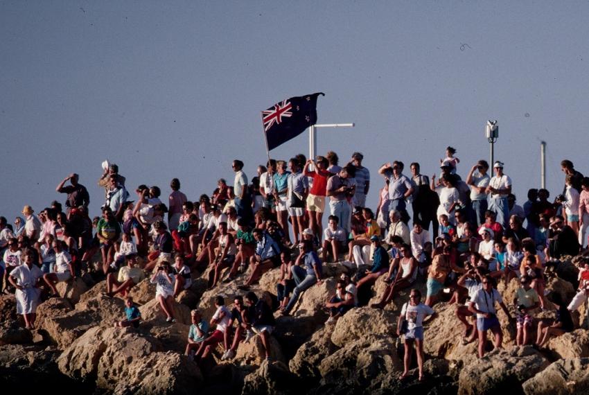 A crowd from 80s era gathers on harbourside rocks with Australian flags in celebration