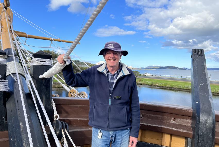 An older man in a dark blue jacket and jeans, and a hat that reads 'Brig Amity' stands on an old sailing vessel and smiles as he holds onto a large white rope. Behind him is a bright blue sky and the sprawling coast of Albany