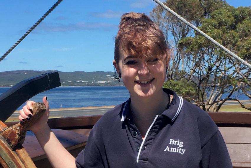 A mid-shot of a young person wearing a navy polo shirt with the words 'Brig Amity' on it smiles at the camera as they hold the steering wheel of the Brig Amity. Behind them, a clear blue sky and the sprawling coastline of Albany is visible