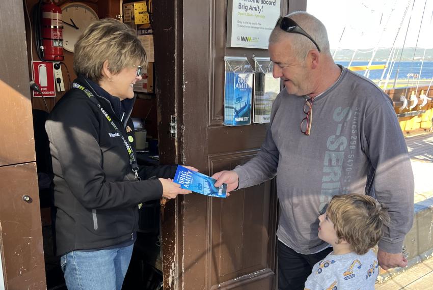 A volunteer with short brown hair and glasses, wearing a black Volunteer shirt and lanyard smiles while handing a blue pamphlet about the Brig Amity vessel to an older man and a young boy. 