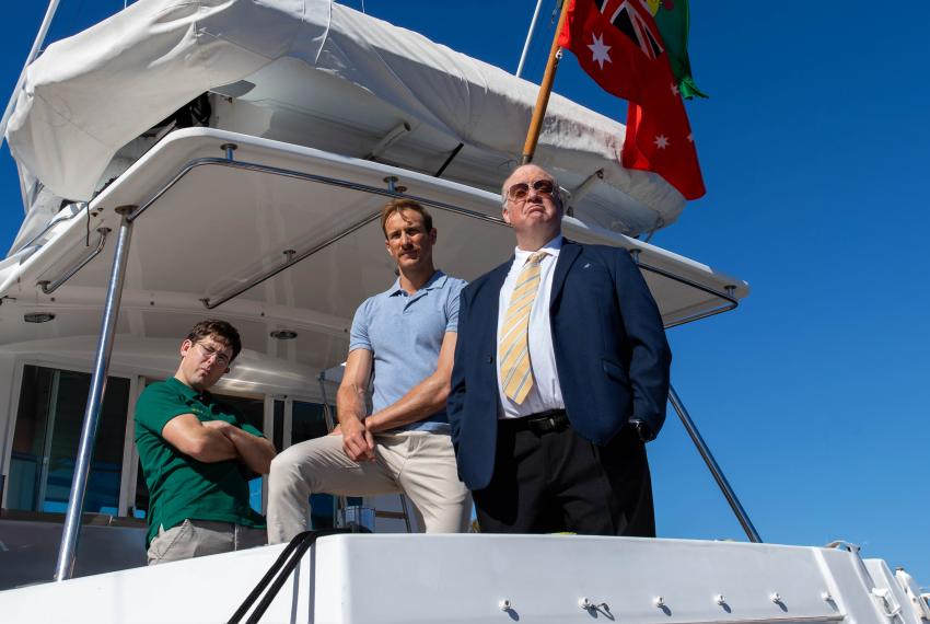 Three men, one dressed in a dark blue suit with a yellow tie and tinted glasses, and the other two in khaki slack pants and a blue and green polo shirt stand on a white boat and stare imposingly into the camera.