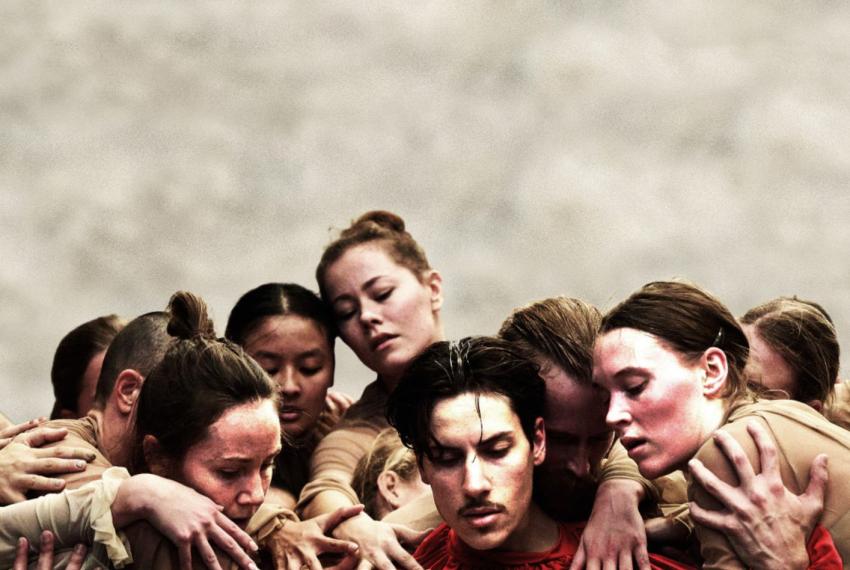A group of dancers in brown textural floaty costumes gather around the main dancer who is dressed in a bright red costumefo the same design. The dancers hand are all interwoven as they clutch at each other creating a mass of bodies.