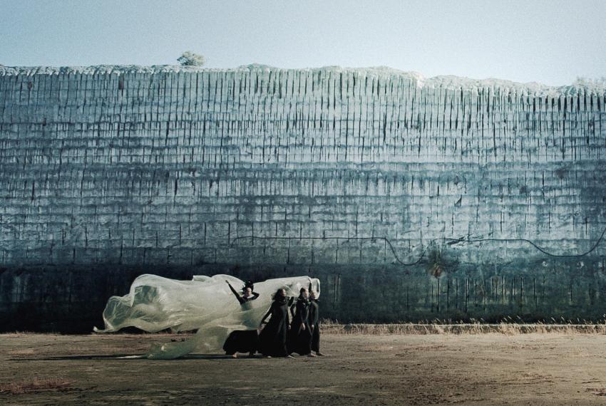 Four dancers wearing long black dresses walk in a procession across a barren space in front of a high blue concrete wall. The dancers hold aloft a large white piece of fabric billowing out behind them in the breeze.