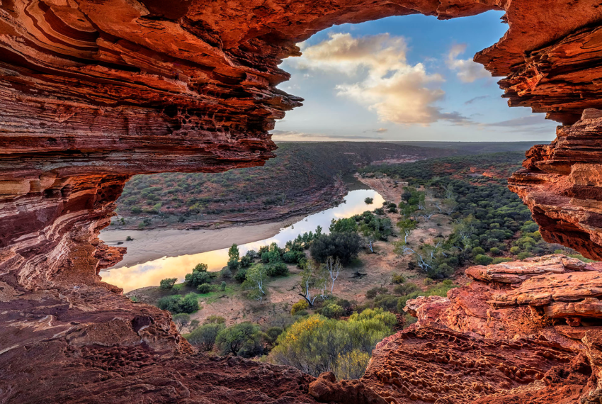 A photograph of Nature's Window, a rock-formation in Kalbarri, with the camera peeing through a colourful banded rock hole looking out over a green and brown landscape and blue sky