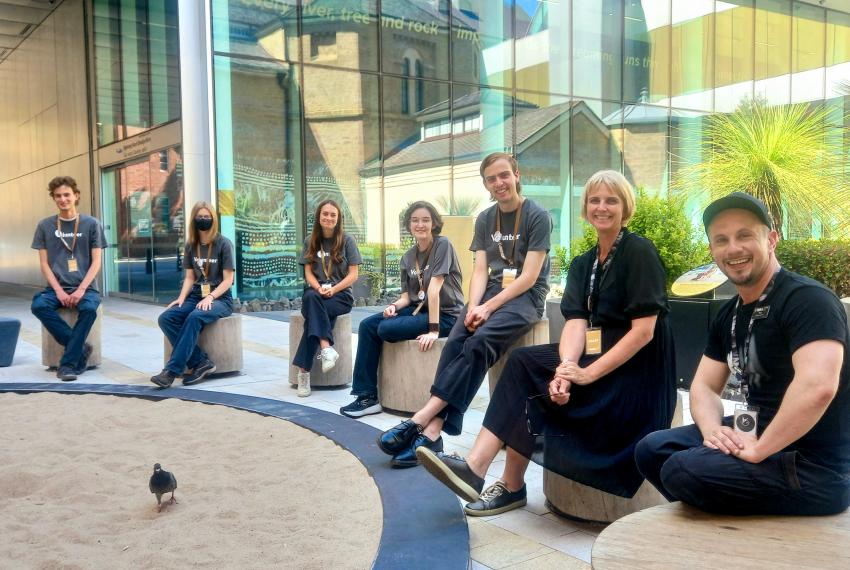 A group of people in grey volunteering shirts and black uniforms sit on individual wooden seats around a sand-filled dance circle in the City Room outside the WA Museum