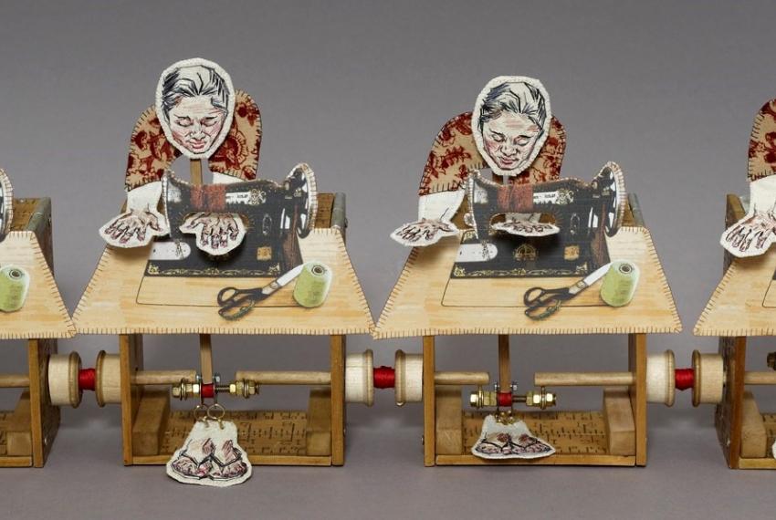 An intricately-crafted series of four paper people perched at tiny wooden and paper sewing machines carefully running something through the machines. The machines appear to be connected by tiny mechanisms as though being able to move when wound