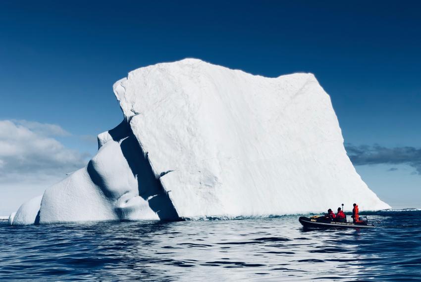 A large white iceberg is framed against a brilliant blue sky and clear rippling blue water, with a tiny boat of researchers at the bottom right corner of the ice berg showing its sheer scale