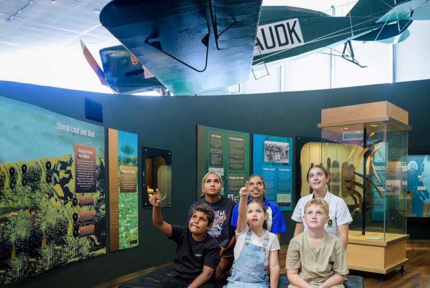 Six young school children sit in the Geraldton Museum surrounded by exhibition panels and showcases look upwards in amazement at a large plane suspended from the ceiling