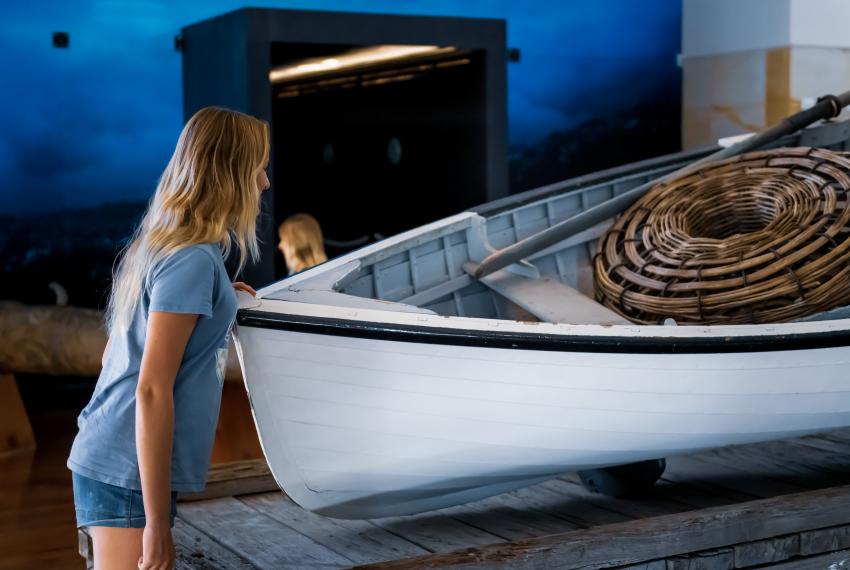 A person with blonde hair and a blue t-shirt with denim shorts leans gently over a white wooden rowing boat to look inside at the large cray net and oar on display at the Museum of Geraldton