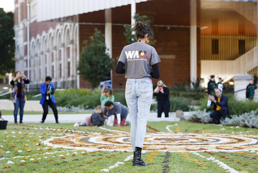 A person wearing a grey shirt with the WA Museum logo and Aboriginal flag on the back walks towards a colourful Mandela-like pattern of coloured sand laid out in the grass in front of the WA Museum