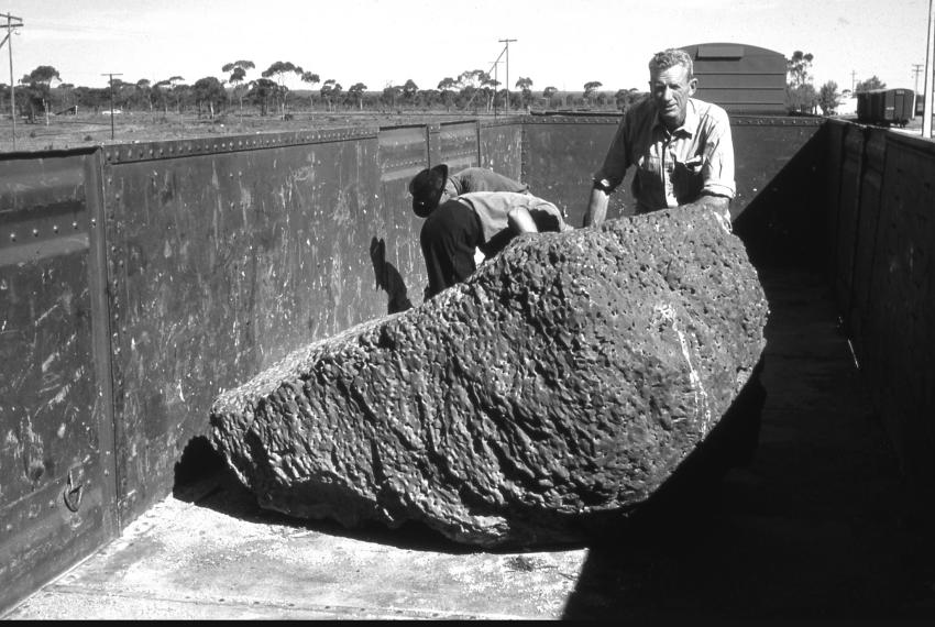 A historic black and white photograph of three men in workwear standing in a large train carriage by a very large piece of meteorite