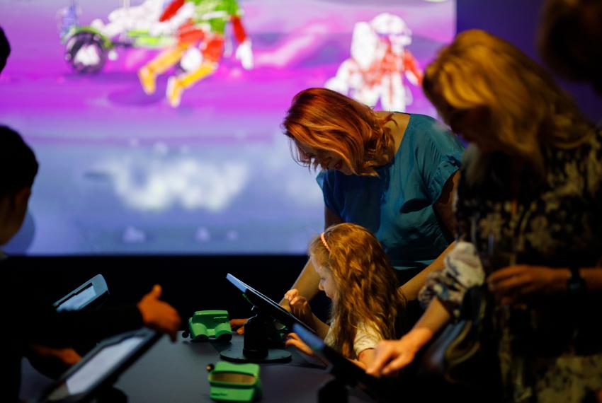 A parent leans over a child using an iPad in a Museum gallery in front of a large screen showing coloured-in astronaut drawings.