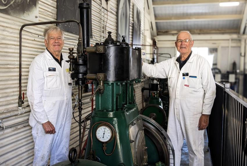 Two older Museum volunteers in white overalls stand on either side of a large green and black steam engine machine and smile at the camera 