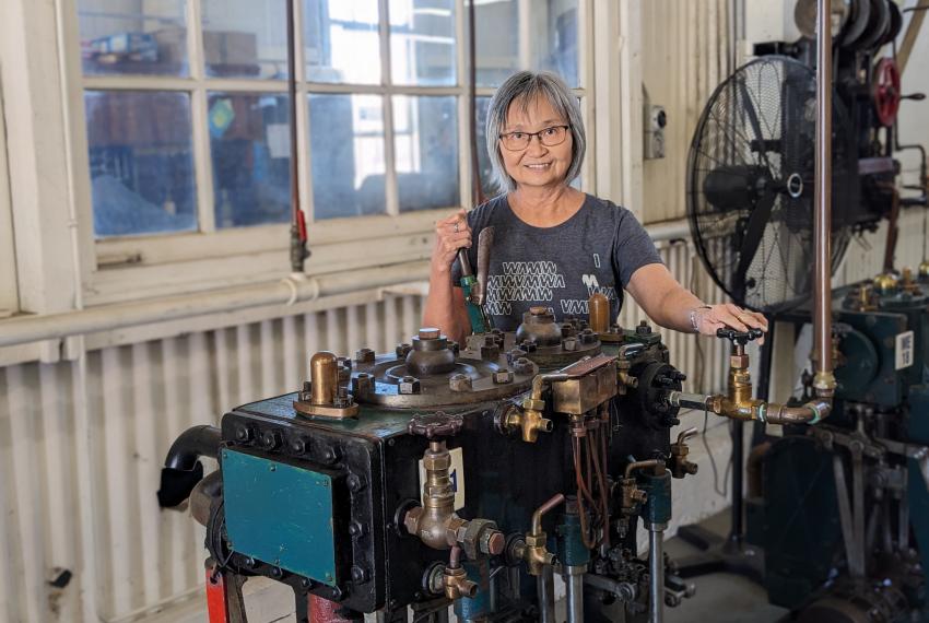 A person with short grey hair and wireframe glasses wears a grey volunteering t-shirt and stands behind a piece of machinery in a workshop setting.