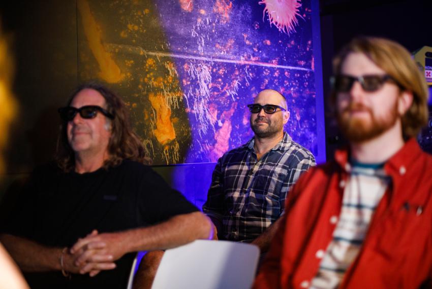 Three individuals sit in a dramatically-lit purple and orange theatrette space and wear black framed 3-d glasses. They stare at something off camera.