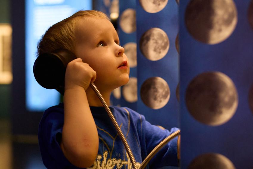 A small child listens to an audio device and looks up to a lunar backdrop