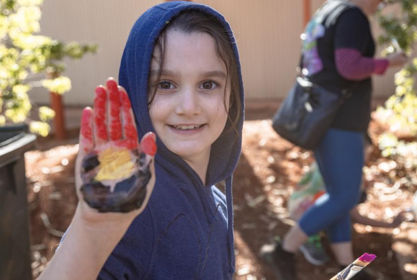 A young child in a blue hoodie smiles into the camera and holds up their hand which is painted with the red yellow and black design of the Aboriginal and Torres Strait Islander flag.