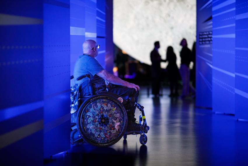 A person in a grey shirt and black glasses with a bald head sits in a wheel chair with an intricately decorated wheel featuring drawings of Australian birds and foliage. They are seated in a vividly-lit blue hallway with a futuristic design in front of a large moon sculpture