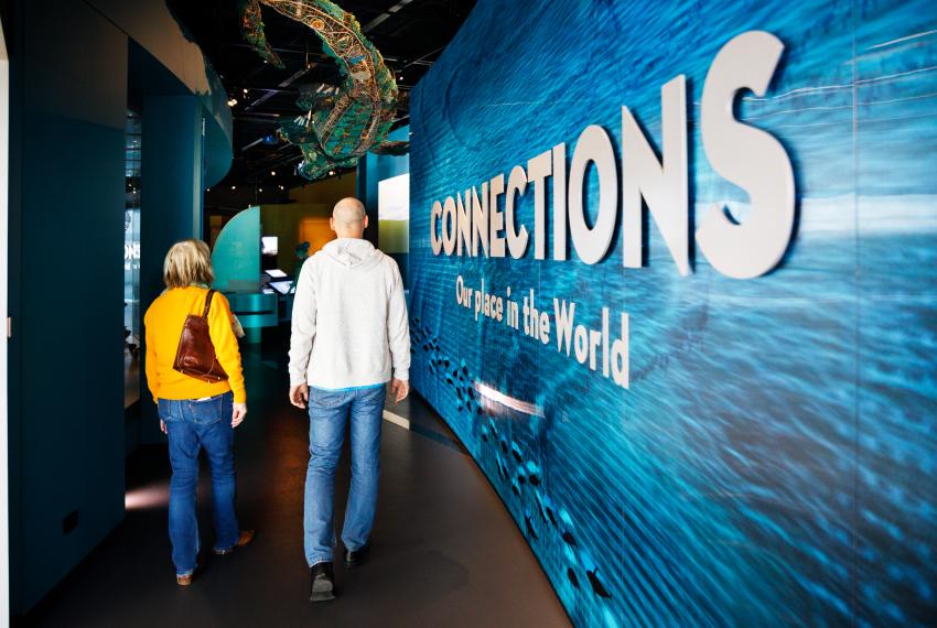  Two people walk through a museum exhibit with a large blue wall displaying "CONNECTIONS Our place in the World."