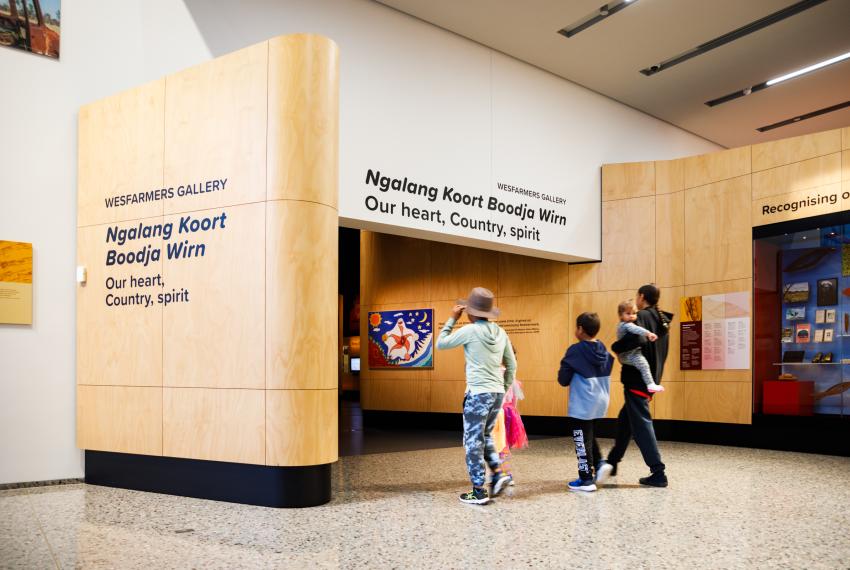 Entrance to Wesfarmers Gallery with wood paneling and text 'Ngalang Koort Boodja Wirn: Our heart, Country, spirit.' Five visitors approach the entrance.