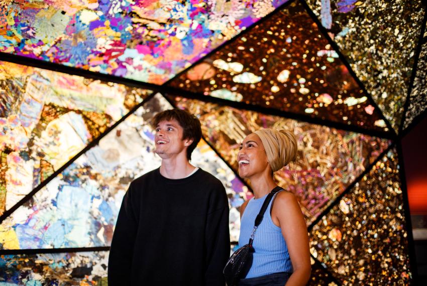 Two people smiling and looking upwards in a colorful, geometric, and illuminated space with intricate patterns.