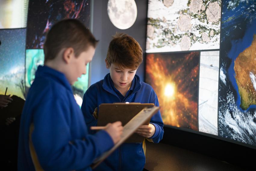 : Two young children in blue school uniform jackets taking notes on clipboards. They are in front of a wall displaying various scientific images, including the moon, a nebula, and Earth