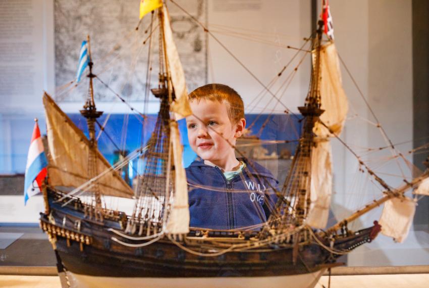 A young child in a blue jumper looks at a detailed ship model in a glass case at a museum exhibit.