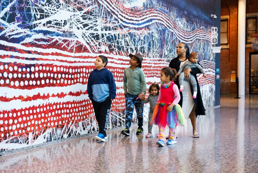 An adult and five children dressed in variously colourful clothes walk indoors next to a colorful, blue and red abstract mural.