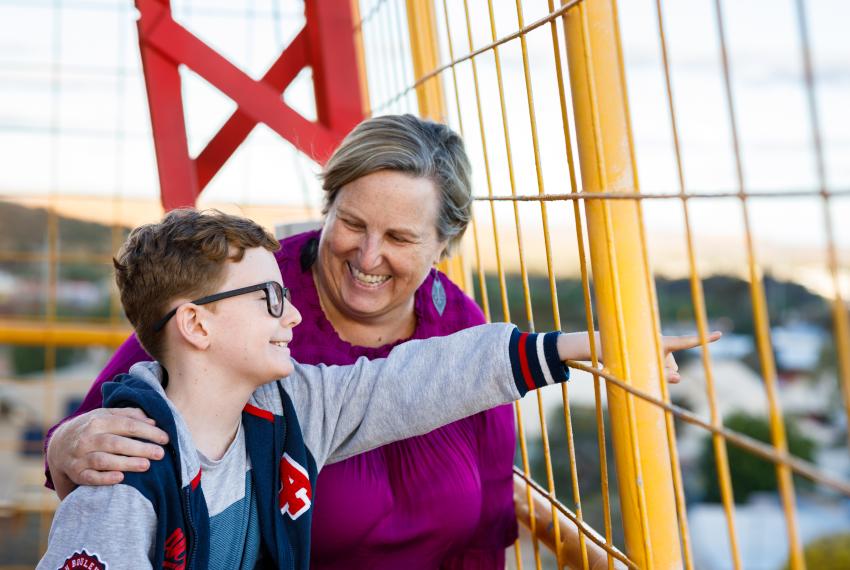 An adult in a purple top embraces a child in a grey and blue hoodie who smiles and points through yellow metal bars towards something in the distance