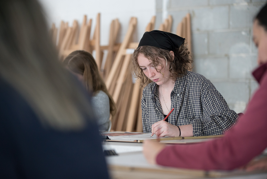 Person drawing with a pencil in a classroom, surrounded by others.