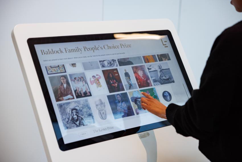 A person interacts with a touchscreen displaying a gallery of artworks titled "Baldock Family People's Choice Prize."