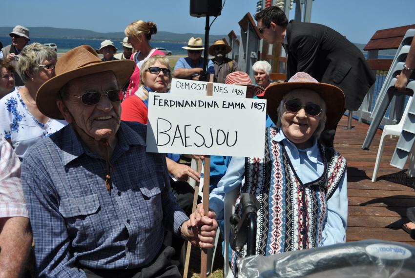 Two individuals sitting outdoors, holding a sign with text, surrounded by other people, beside a body of water.