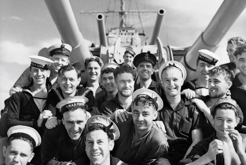 group of men in navy uniform on HMAS Perth smiling for a photo sitting under the ships guns