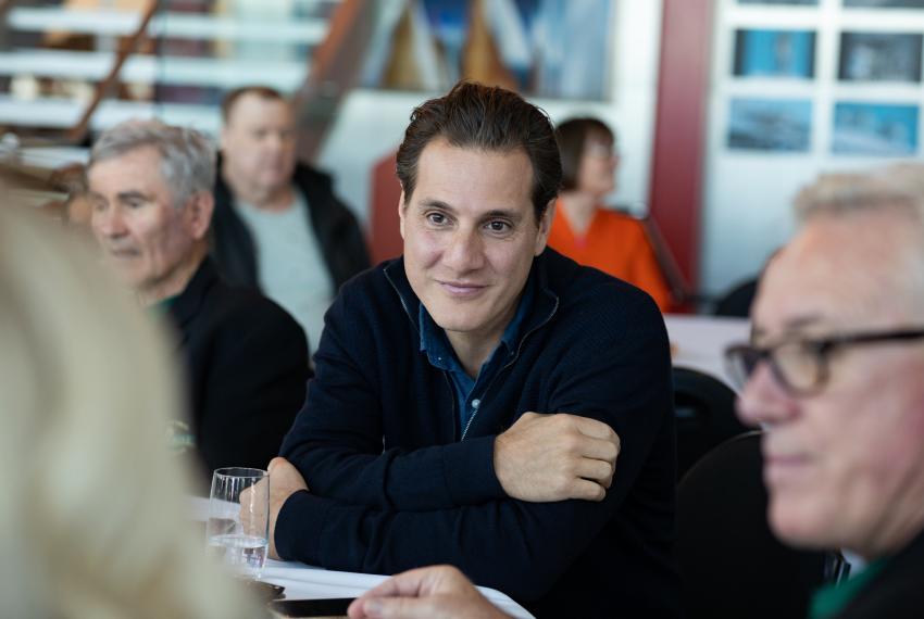 A person in a navy sweater sits at a table, smiling at the camera, with others in a blurred background.