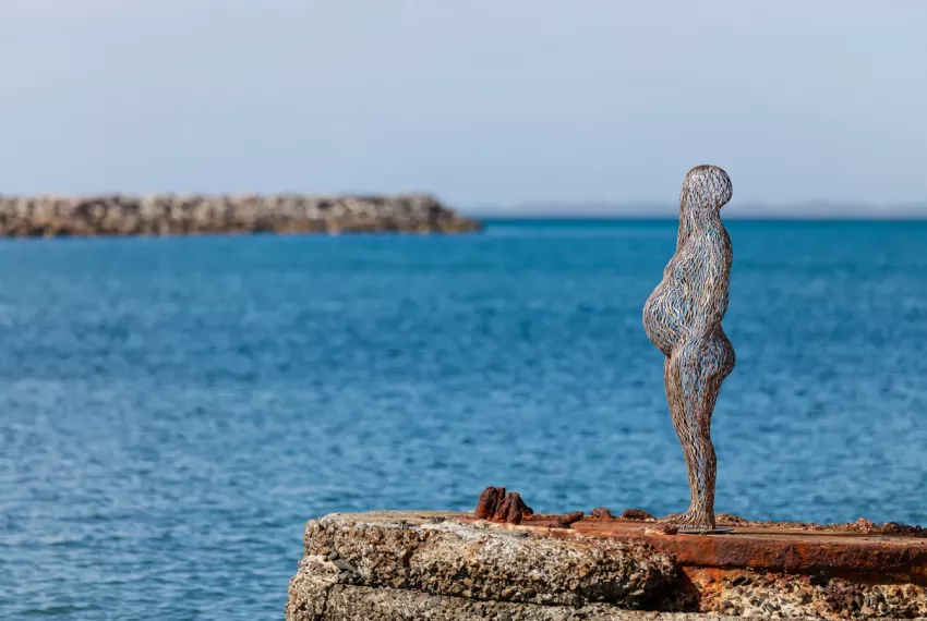 A metal sculpture of pregnant person positioned on a red rusted plinth in the ocean with vibrant blue water visible around and behind it