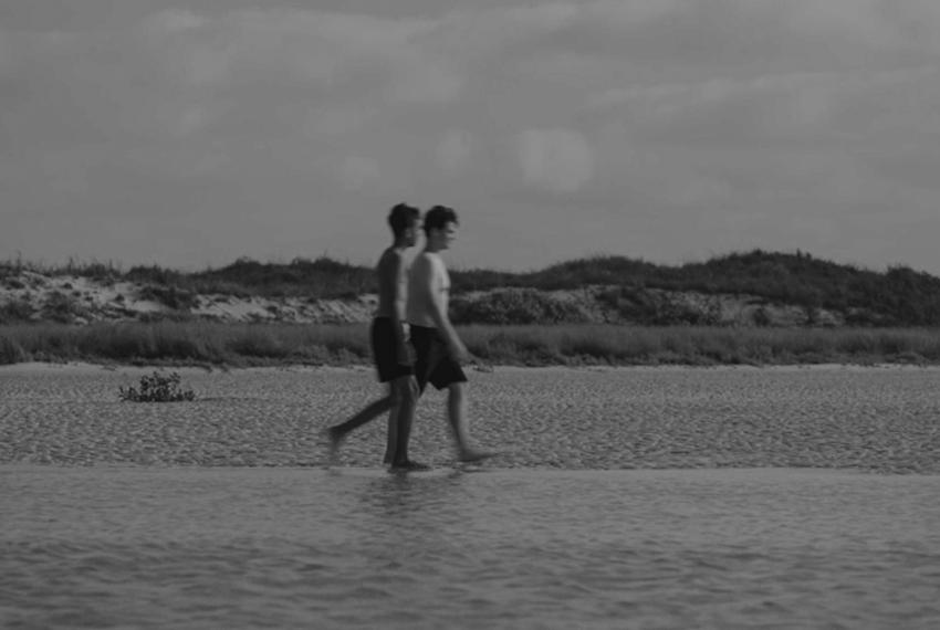 Black and white still of two boys walking across a beach