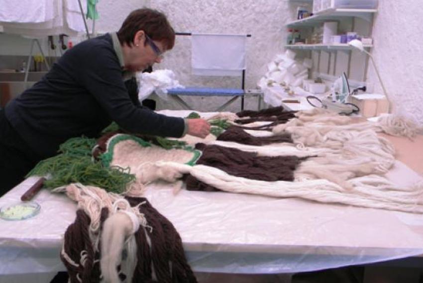 Person working on a textile project with yarn strands of green, brown, and cream on a table in a craft workspace.