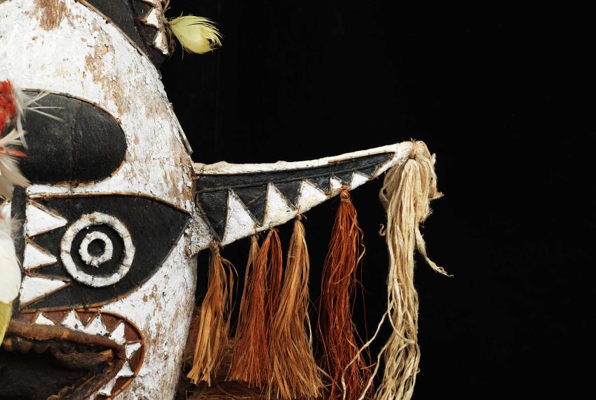 Close-up of a tribal mask with black, white, and beige patterns, featuring circular and triangular designs, tassels, and feathers.