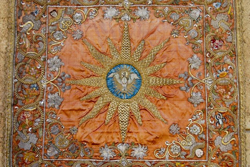 An embroidered textile with a central pelican emblem and sunburst on an orange background, surrounded by floral motifs.