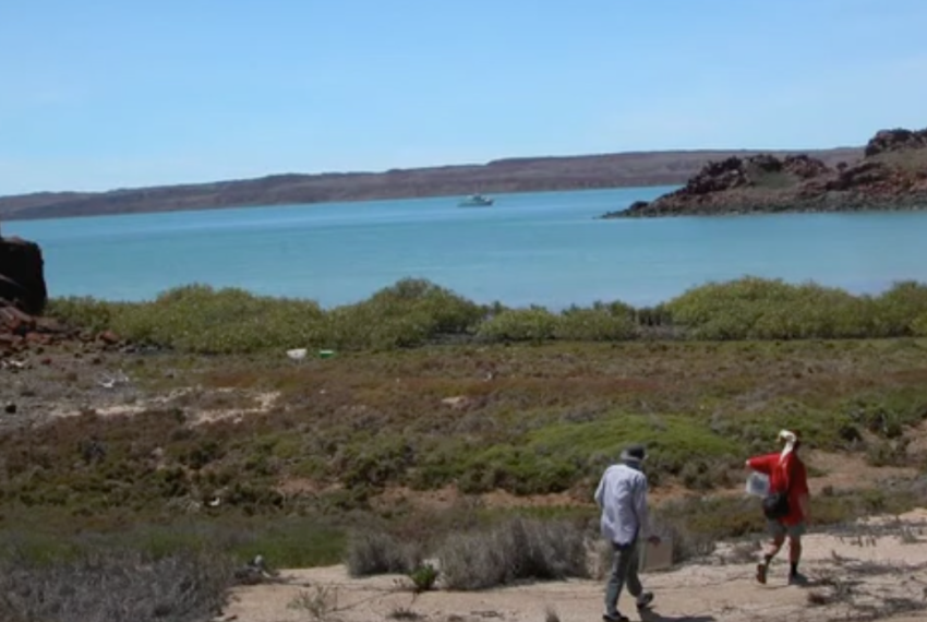 Two people walking near a rocky shore with turquoise water and a distant peninsula.