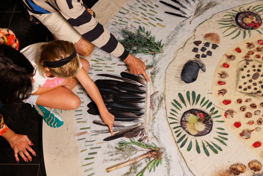 Aerial photograph of three people admiring a large rug decorated with natural materials like rocks, feathers and leaves