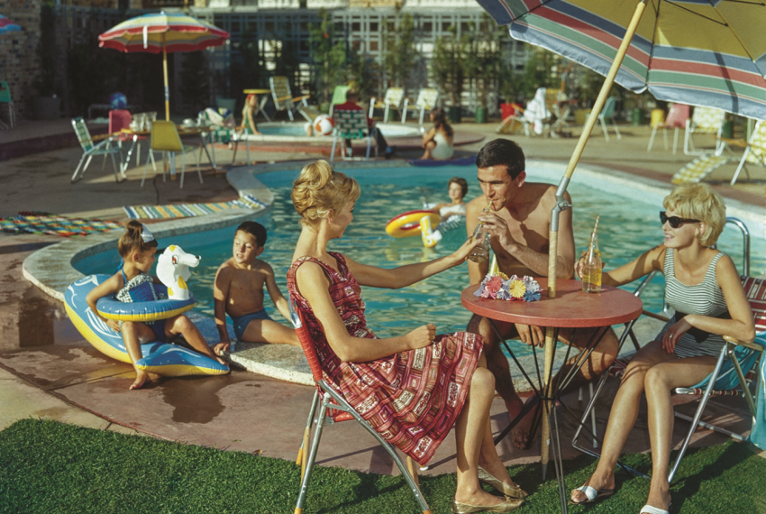 A vintage coloured photo of three people sitting under a pool umbrella at a small round table while their kids play in a motel pool behind them.