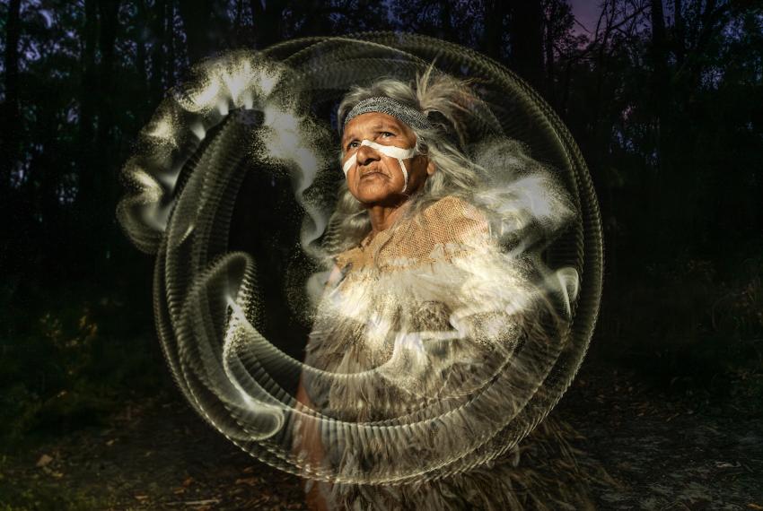 an Aboriginal Elder with traditional ochre on her face and arms looks up. She wears clothing made of emu feathers. There is a white circular halo and dust around her