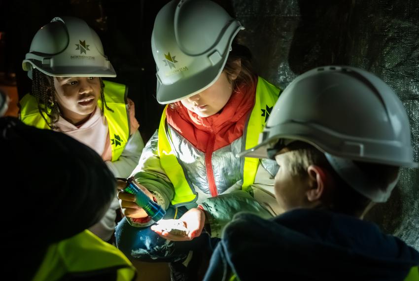 Children wearing hard hats and safety vests, examining an object in dim light.