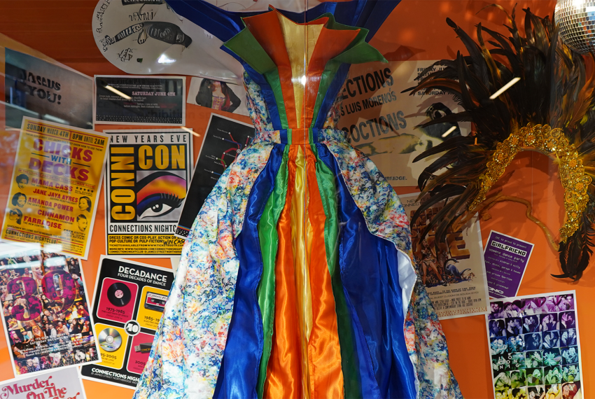 A colourful display featuring a multicoloured dress, a feather headdress, and various vibrant posters against an orange background.