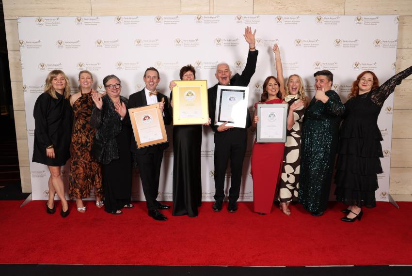 A happy group of 10 people from the WA Museum holding award certificates Infront of the Tourism award media wall after receiving an award