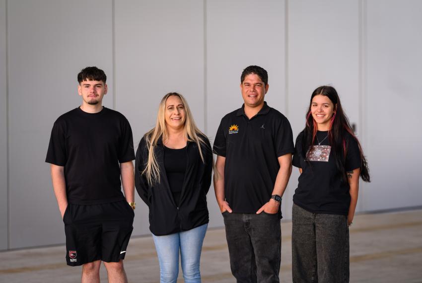 Four individuals standing side by side against a light grey wall, all wearing dark clothing. The group is positioned in a casual stance, with hands either by their sides or in pockets