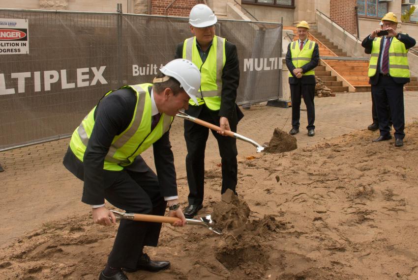 Premier McGowan bending with a ceremonial shovel and Minister Templeman standing to his left.  They are turning sod by digging the soil at the site of the New Museum.  Both are wearing fluro yellow vests and hard hats.